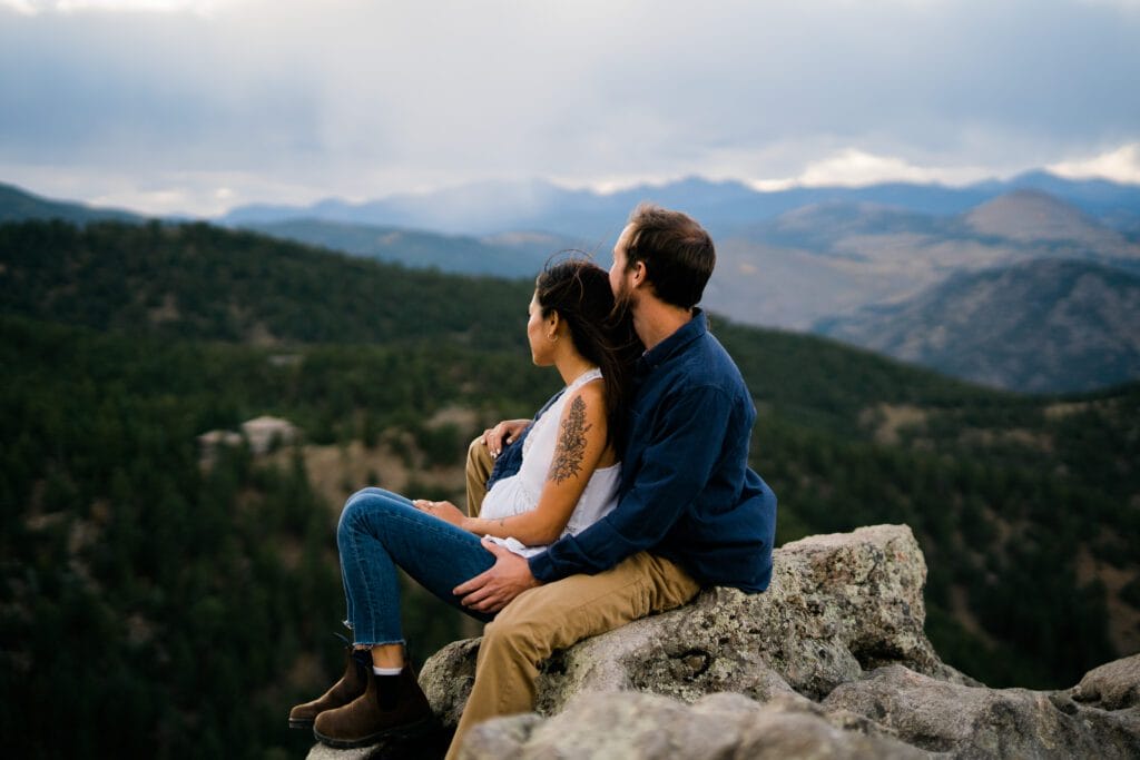 A couple sits on a rocky ledge, overlooking a vast, tree-covered mountain landscape under a cloudy sky. The woman leans against the man as they gaze into the distance, both dressed casually in jeans and boots.