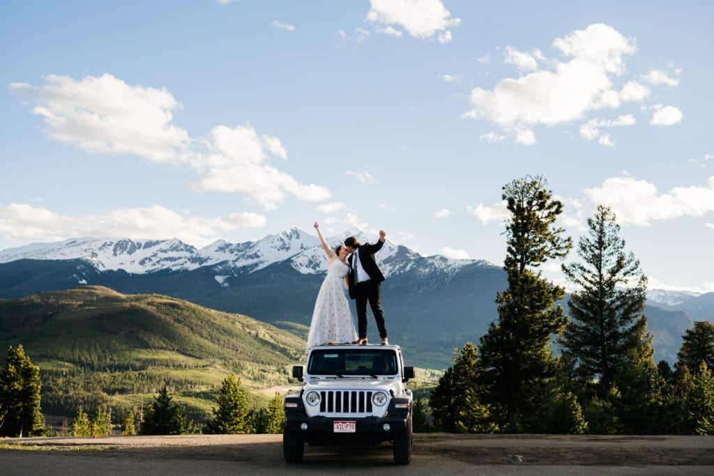 A bride and groom stand on top of a white Jeep, raising their arms joyfully, with a scenic backdrop of mountains, trees, and a partly cloudy sky.