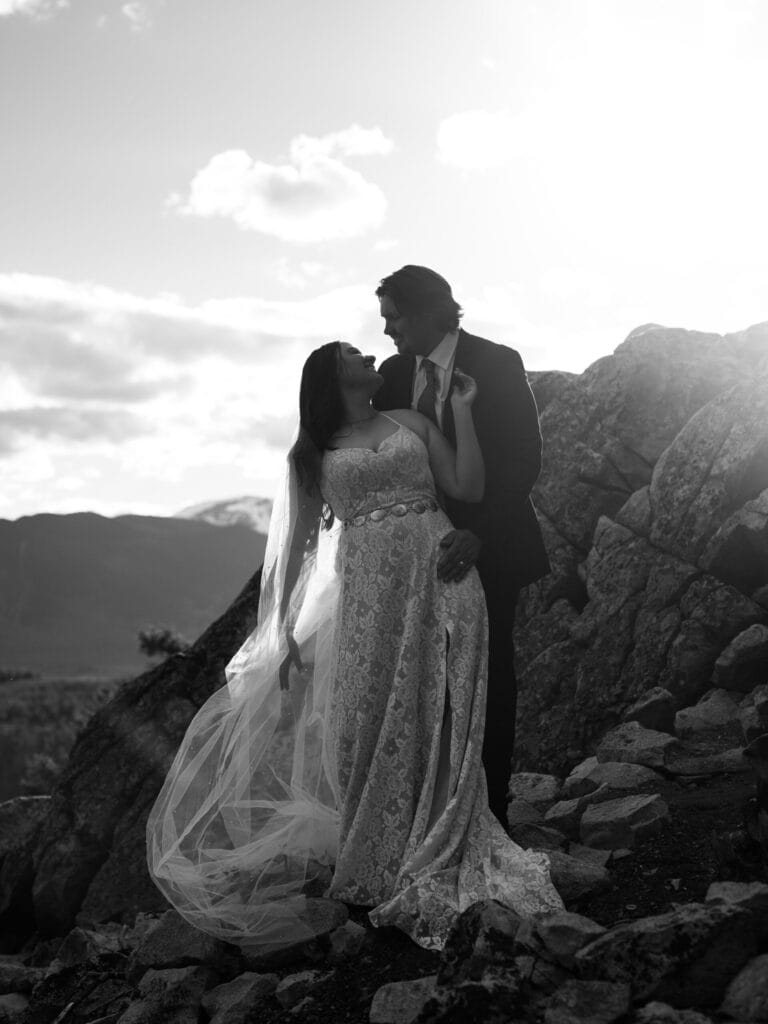 A bride and groom stand on rocky terrain, gazing at each other lovingly. The bride wears a lace gown with a long veil, and the groom is in a suit. Sunlight streams behind them, creating a romantic, dramatic scene.