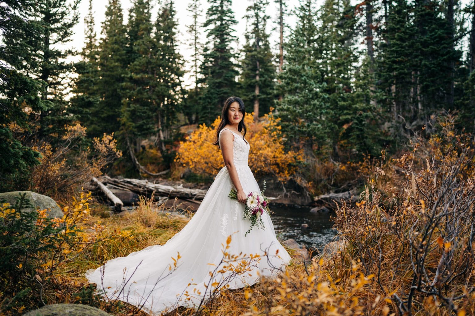A bride in a white gown holding a bouquet stands outdoors by a stream, surrounded by autumn trees and foliage, with tall evergreen trees in the background.during her Brainard Lake Elopment