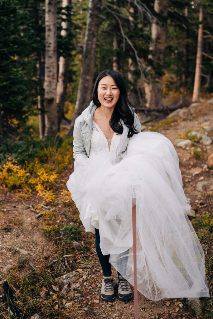 A woman in a white wedding dress and grey jacket stands outdoors on a forested trail, smiling brightly. She holds up her dress, revealing hiking shoes, blending elegance and adventure.