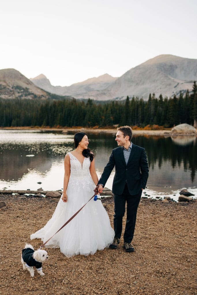 A bride and groom hold hands and smile at each other while walking a small dog in a tuxedo near a lake, with mountains and pine trees in the background.