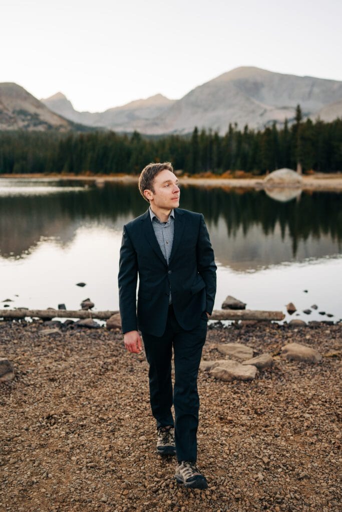 A man in a dark suit and hiking boots walks on rocky ground beside a lake, with pine trees and mountains in the background.