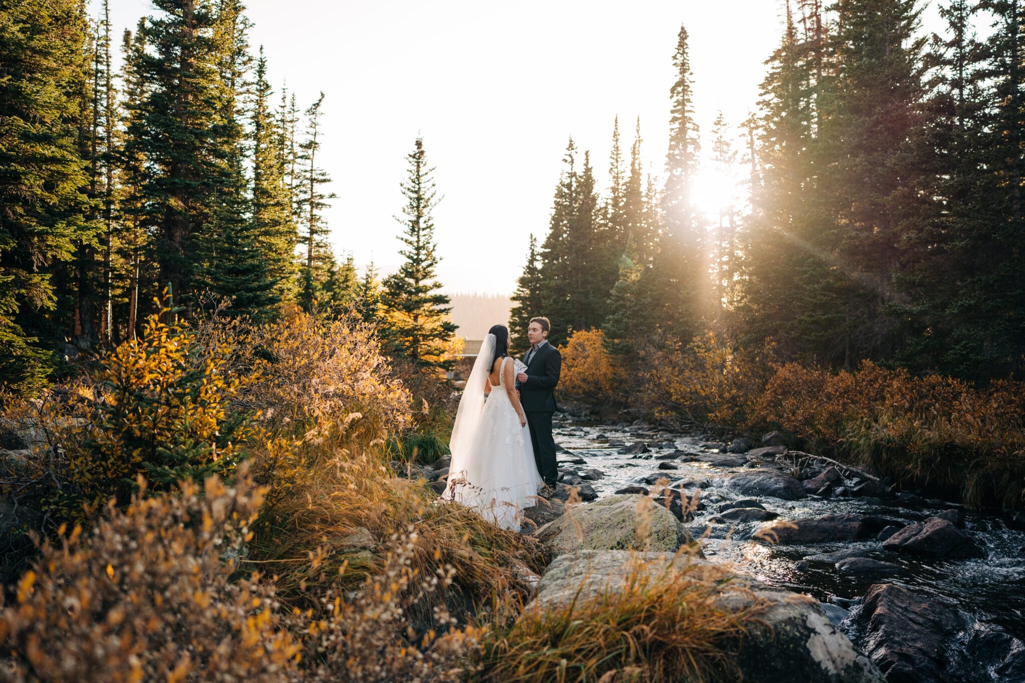 A bride and groom stand together beside a rocky creek at Brainard Lake, surrounded by tall evergreen trees and autumn foliage, with sunlight streaming through the forest—perfect inspiration for an elopement.