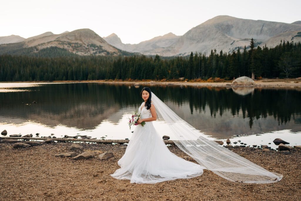 A bride in a white wedding gown and long veil stands by a calm mountain lake, holding a bouquet, with pine trees and mountains in the background under a clear sky.