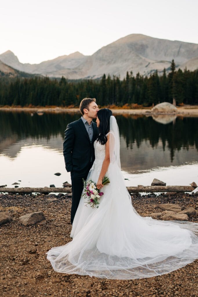 A groom in a dark suit kisses a bride in a white gown and veil on the forehead as they stand by a calm lake, surrounded by trees and mountains at sunset. The bride holds a bouquet of flowers.