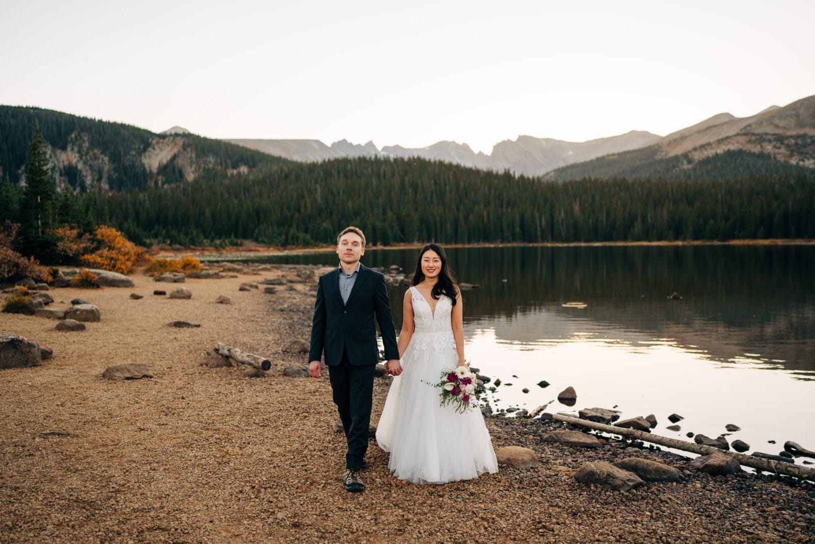 A bride and groom walk hand-in-hand along a rocky lakeshore, surrounded by mountains and pine trees, with calm water reflecting the landscape at sunset. The bride holds a bouquet and wears a white gown; the groom is in a dark suit.
