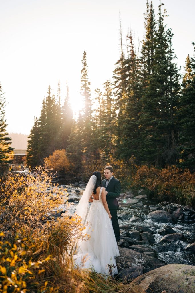 A bride and groom stand on rocks by a flowing creek, surrounded by tall pine trees and autumn foliage, with sunlight shining through the trees in the background during their elopement in telluride.