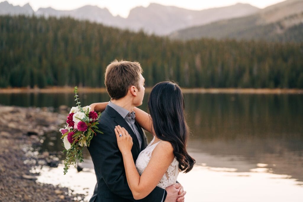 A bride and groom stand by a calm lake, embracing and looking at distant forested mountains. The bride holds a bouquet of pink and white flowers, and they are dressed in wedding attire.