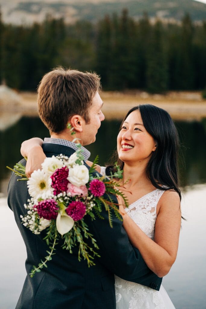 A bride and groom embrace outdoors near a lake, smiling at each other. The bride holds a bouquet of white and pink flowers, and evergreen trees and mountains are visible in the background.