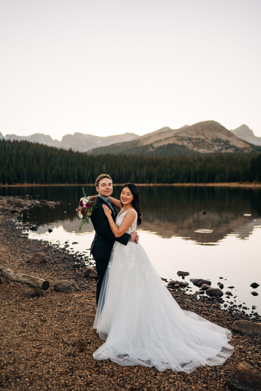 A bride and groom embrace by a calm lake with mountains and pine trees in the background. The bride wears a white gown and the groom holds a bouquet, both smiling in the soft outdoor light.