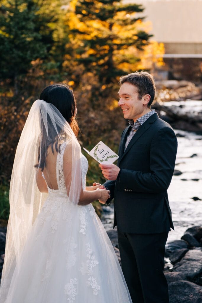 A groom in a dark suit smiles while reading vows to a bride in a white wedding dress and veil. They stand outdoors by a rocky stream with autumn trees in the background.