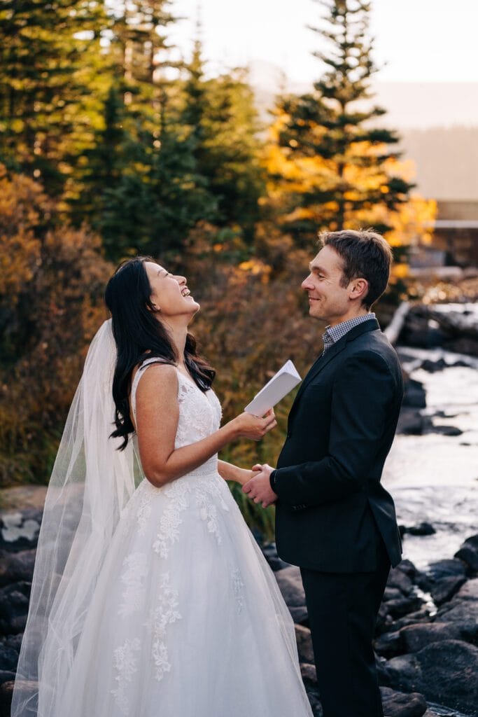 A bride in a white dress and veil laughs while holding hands with the groom in a dark suit by a river, surrounded by trees with autumn foliage during their elopement in Telluride.