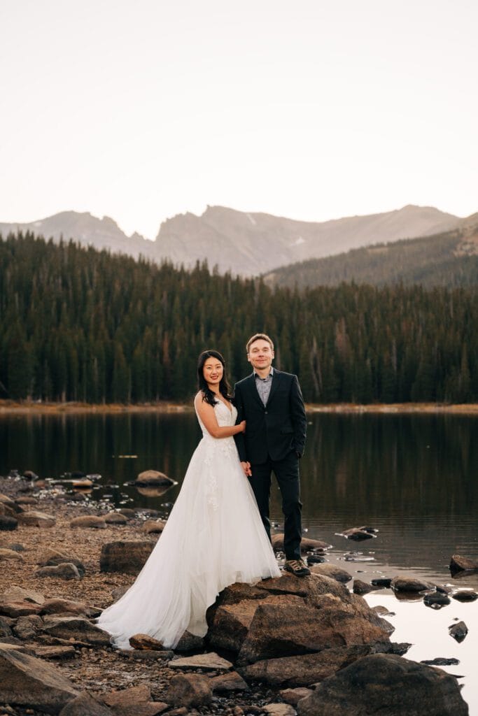 A bride in a white wedding gown and a groom in a dark suit stand smiling on rocks by a calm lake, with pine trees and mountain peaks in the background at sunset.