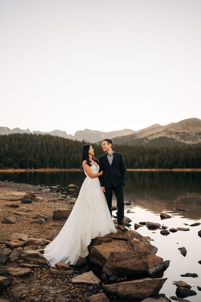 A bride in a white gown and a groom in a dark suit stand on rocky ground by a calm lake, smiling at each other, with pine trees and mountains in the background.