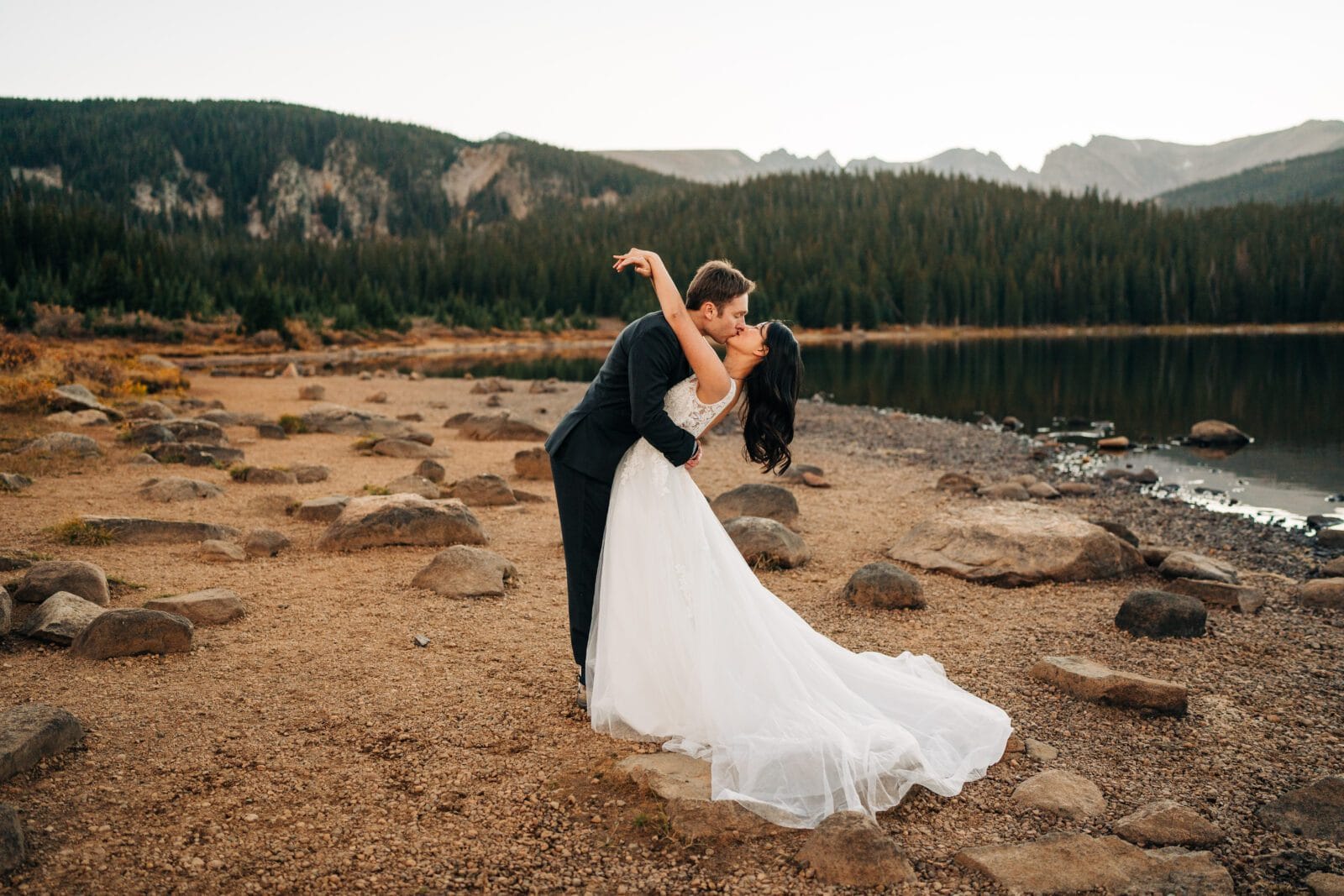 A bride and groom share a romantic kiss as the groom dips the bride on a rocky lakeshore, with pine trees and mountains in the background. The bride wears a flowing white gown; the groom is in a black suit.