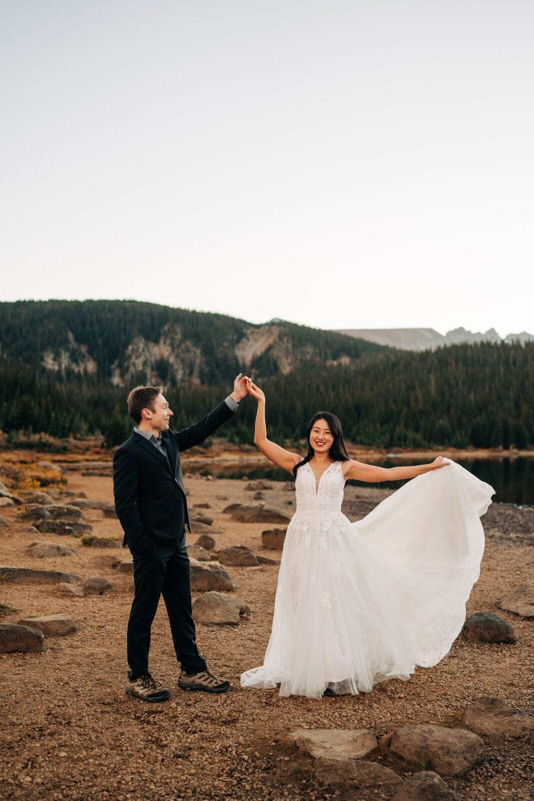 A bride in a white gown holds her dress and twirls with a groom in a black suit on a rocky lakeshore, surrounded by pine trees and mountains under a clear sky.
