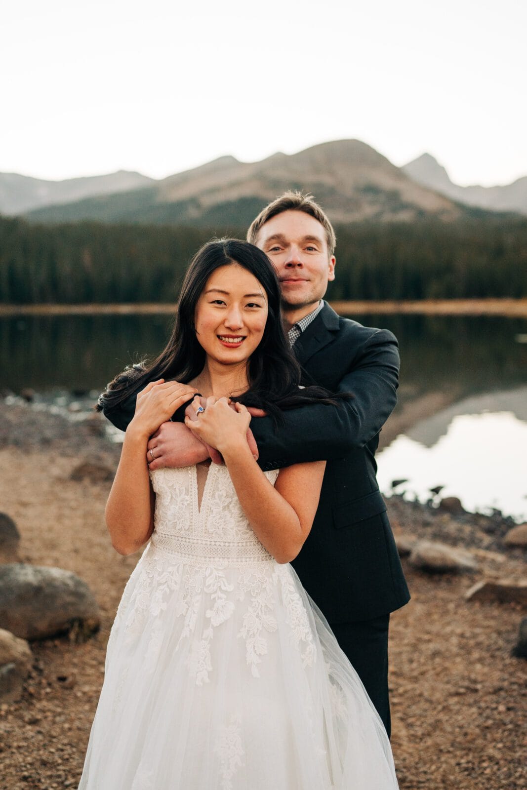 A bride in a white lace dress stands in front of a groom in a dark suit by a lake with mountains in the background. The groom hugs her from behind, both smiling at the camera amidst a natural outdoor setting.