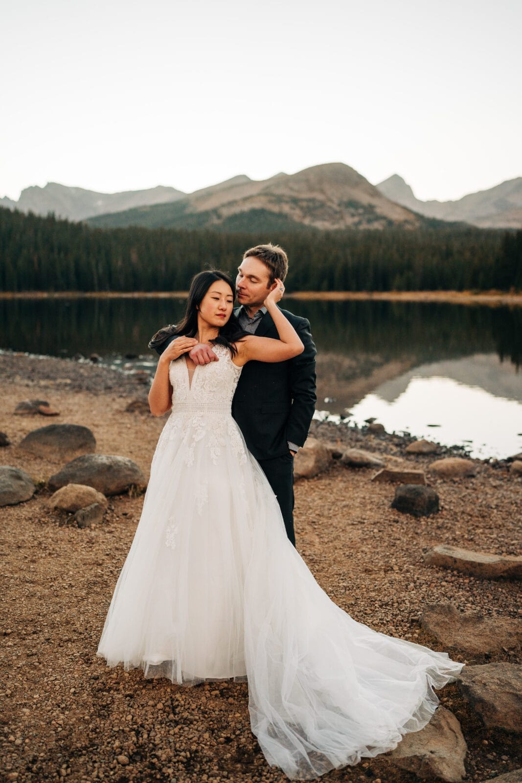 A bride in a white gown and a groom in a dark suit embrace by a calm lake, surrounded by rocks and trees, with mountains in the background at sunset.