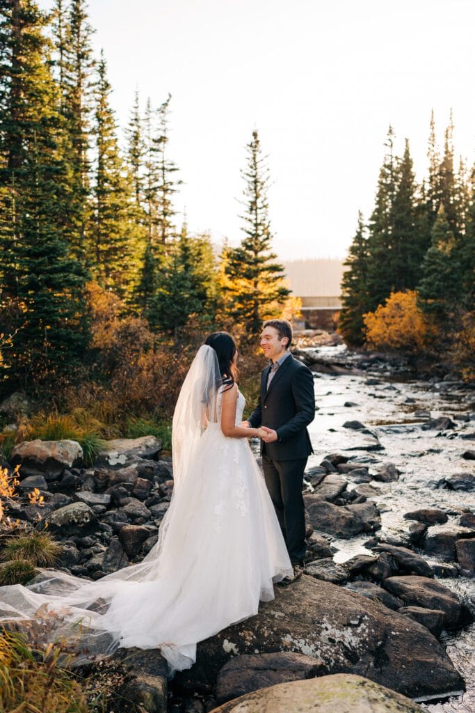 A bride and groom stand on rocks by a stream, holding hands and facing each other, surrounded by evergreen trees and autumn foliage, with sunlight filtering through the forest.