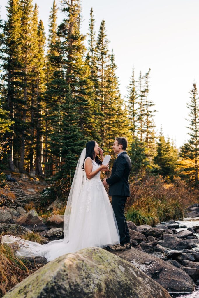 A bride and groom stand on rocks by a stream in a forest, exchanging vows and smiling at each other. The bride wears a white dress and veil, and the groom is in a black suit. Tall pine trees fill the background during their elopement in Telluride.