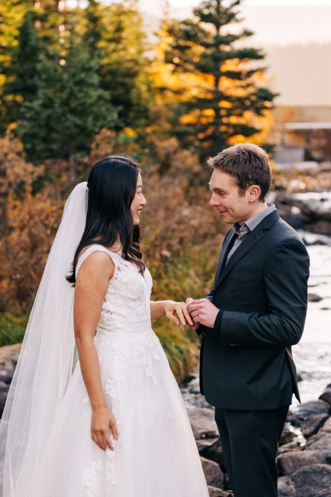 A bride in a white gown and veil smiles at a groom in a dark suit as they stand together outdoors by a rocky stream, with autumn trees in the background.