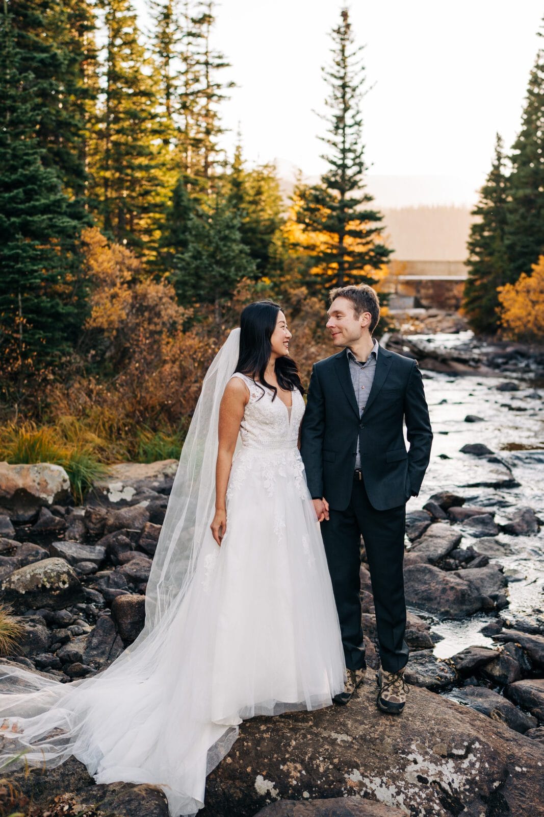 A bride in a white gown and veil stands hand-in-hand with a groom in a suit by a rocky stream surrounded by pine trees and autumn foliage, both smiling at each other.