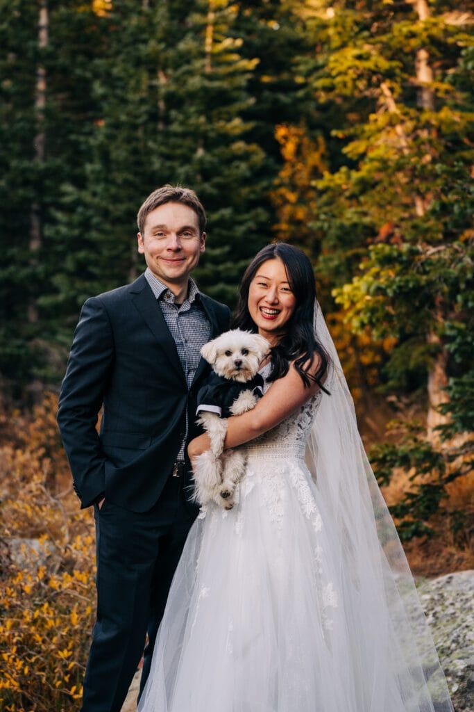 A bride in a white wedding dress and veil stands beside a groom in a black suit, smiling outdoors with trees in the background. The bride holds a small white dog dressed in a black outfit.