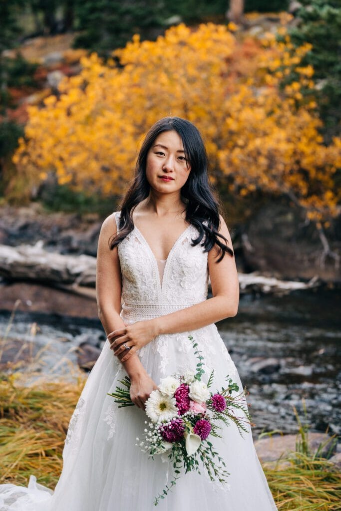 A woman in a white wedding dress holds a bouquet of flowers while standing outdoors near a stream, with yellow autumn foliage in the background.