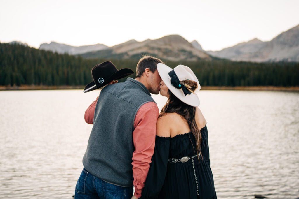 A couple in hats kisses by a lake, with pine trees and mountains in the background at sunset.