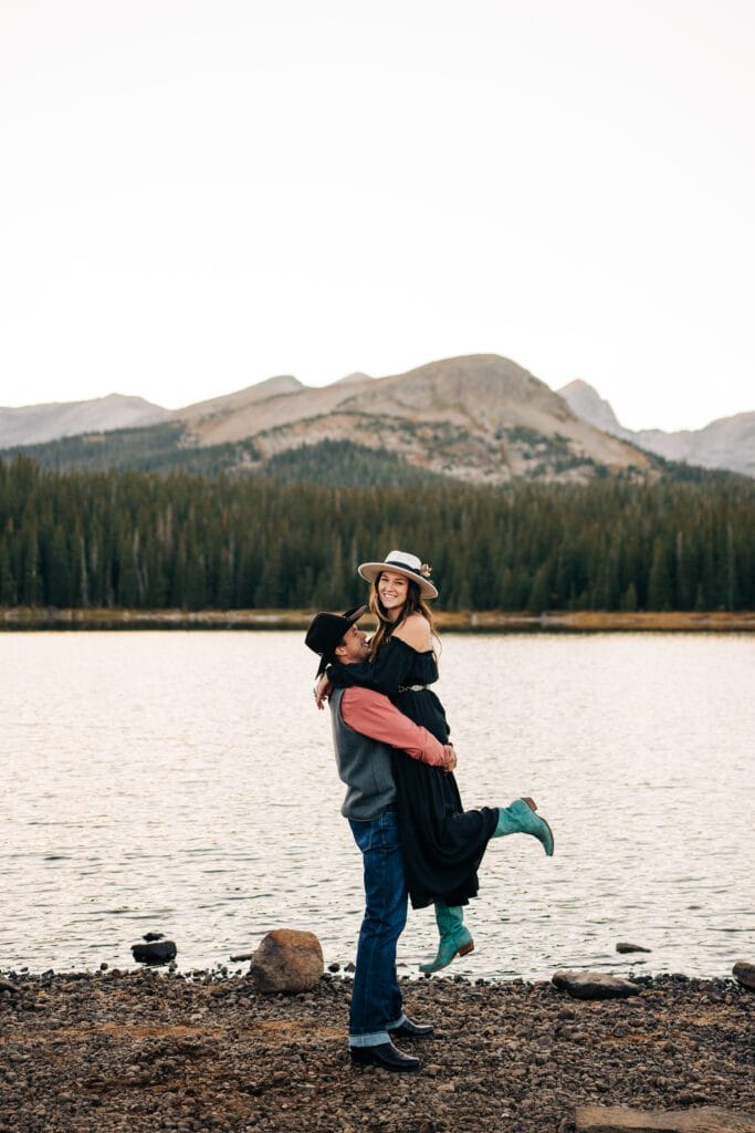 A man lifts a smiling woman by a lake, with mountains and pine trees in the background. The woman wears a hat, a long dress, and turquoise boots. Both appear happy and outdoors in nature.