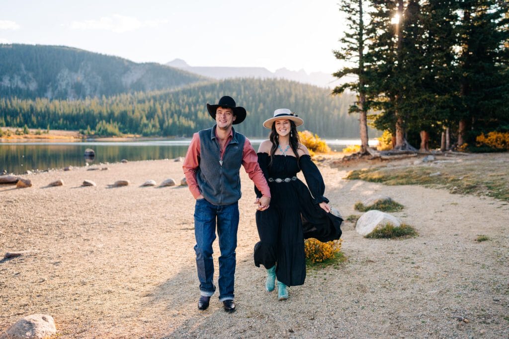 A smiling couple in western-style clothes walk hand in hand along a lakeshore, surrounded by pine trees and mountains, with sunlight shining through the trees.
