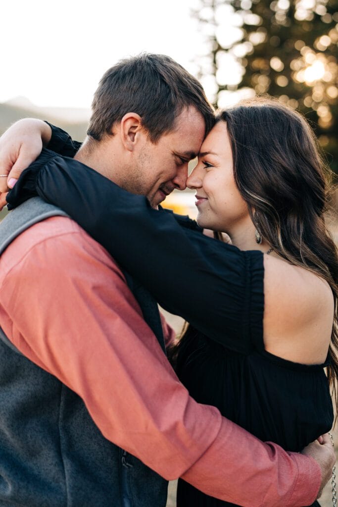 A man and woman embrace closely, touching foreheads and smiling with closed eyes, outdoors with trees and soft sunlight in the background.