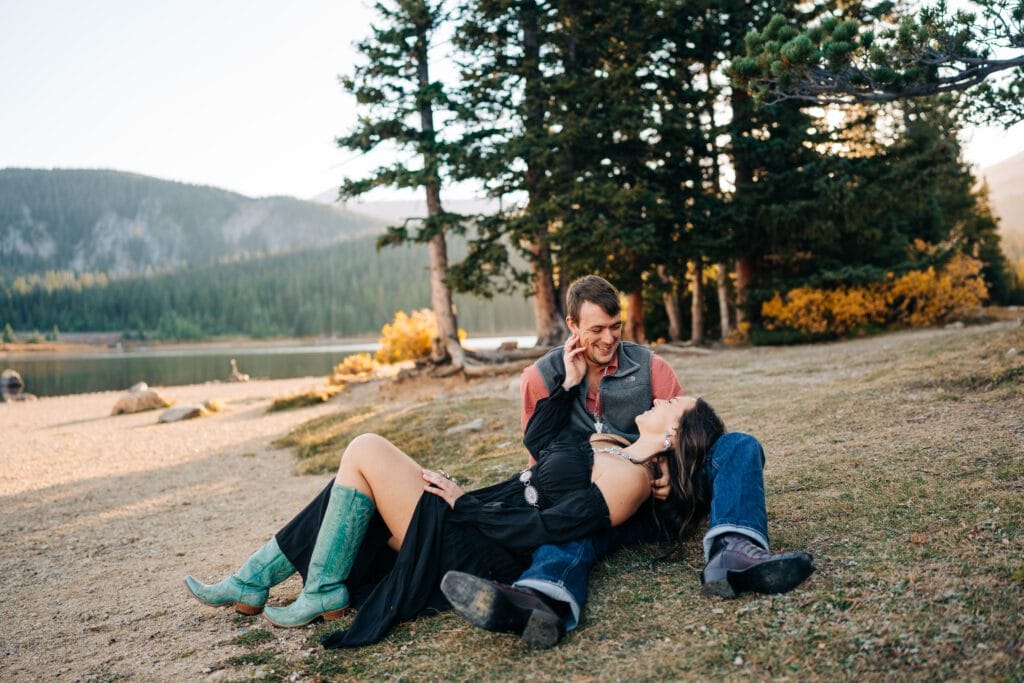 A couple sits on the grass by a lake and pine trees. The woman, wearing a black dress and turquoise boots, lies with her head in the mans lap, both smiling and gazing at each other in a relaxed, outdoor setting.