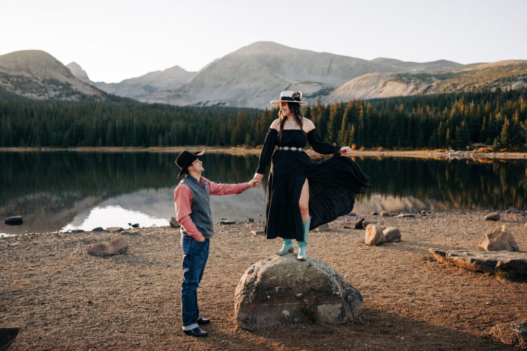A man in jeans and a hat holds hands with a woman in a flowing black dress and hat standing on a rock by a lake, with mountains and trees in the background at sunset.