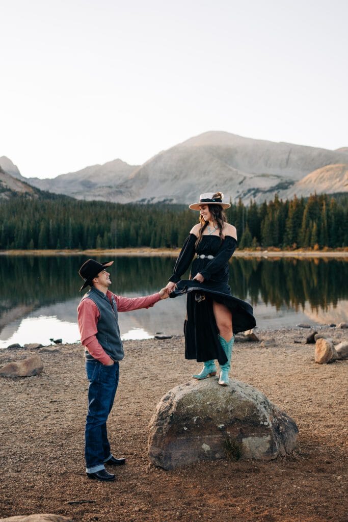 A man in a cowboy hat holds hands with a woman in a dress, turquoise boots, and wide-brimmed hat as she stands on a rock by a lake, with pine trees and mountains in the background at sunset.