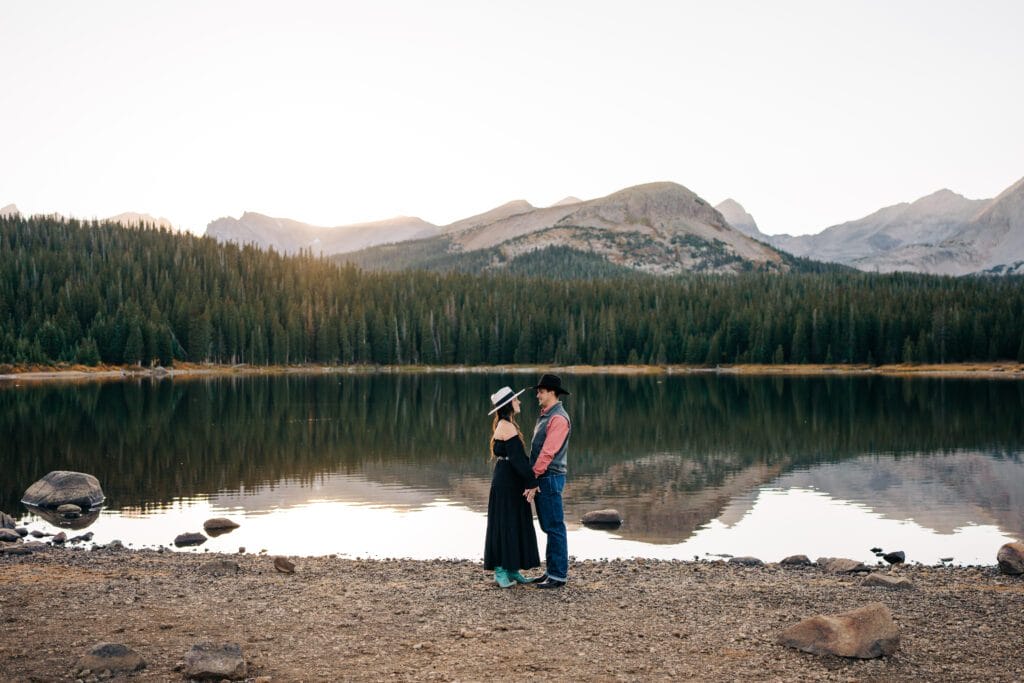A couple stands facing each other by a calm lake, surrounded by pine trees and mountains, with the couple’s reflection visible in the water. The woman wears a dress and hat, and the man wears a cowboy hat and boots.