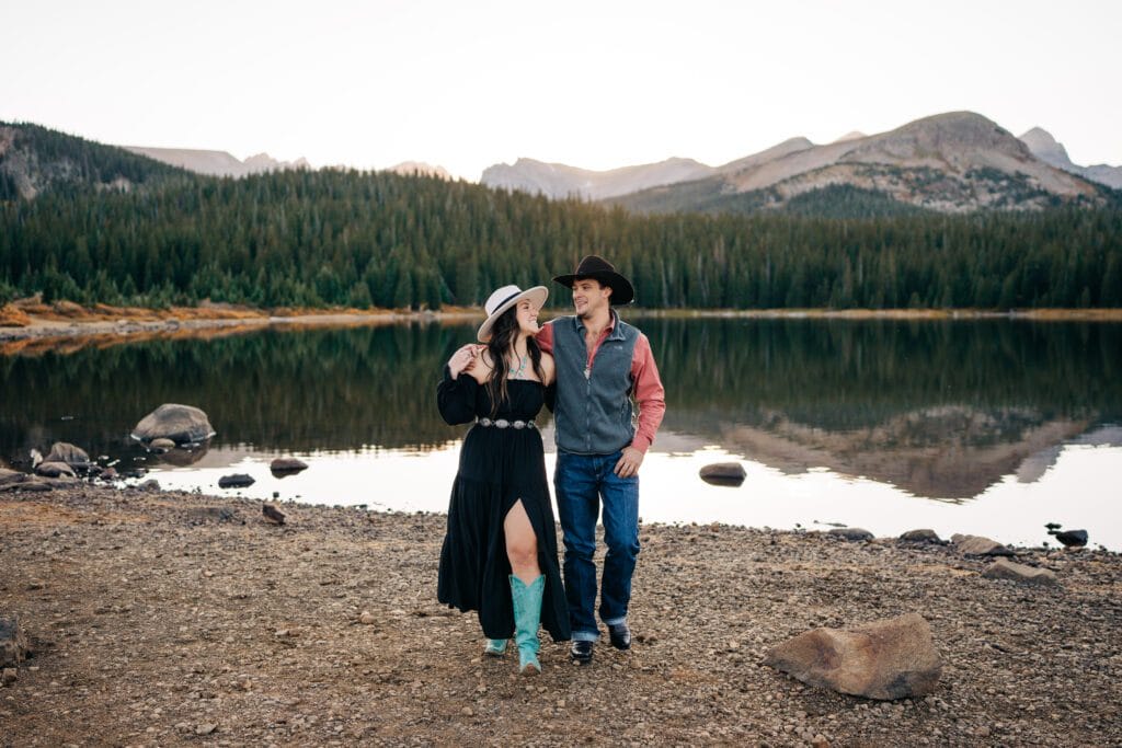A couple dressed in western attire walks arm in arm by a lake with mountains and pine trees in the background, under a clear sky.