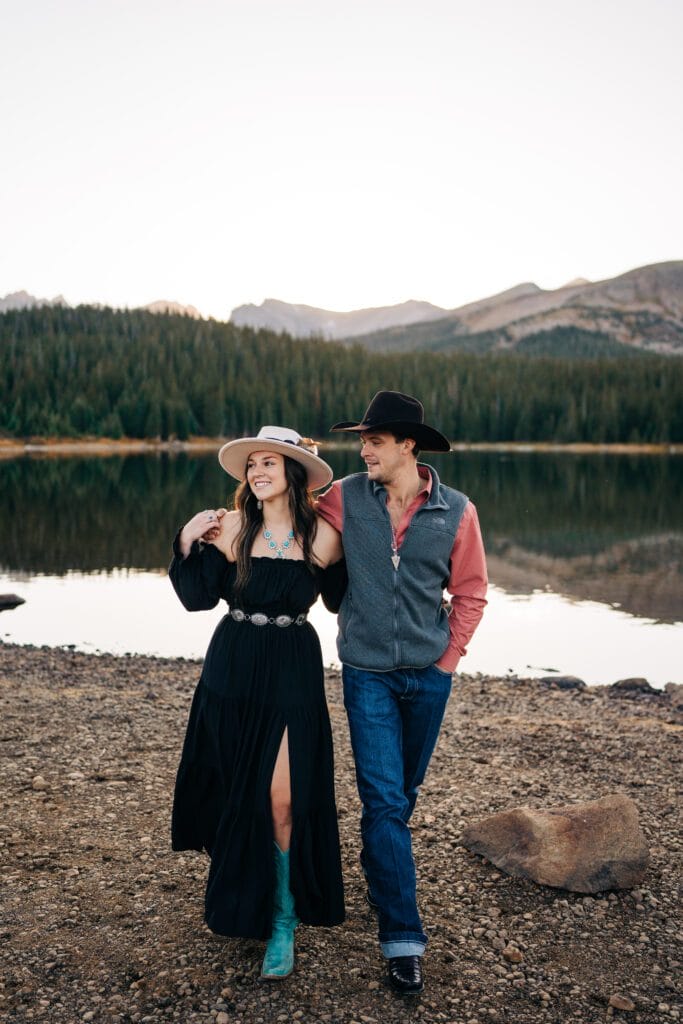 A woman in a black dress and hat walks arm in arm with a man in jeans and a cowboy hat by a lake, with pine trees and mountains in the background.