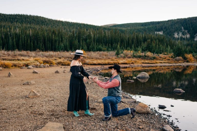 A man in a cowboy hat kneels by a lakeshore, proposing to a woman in a black dress and hat. They are surrounded by trees and mountains in the background under a clear sky.