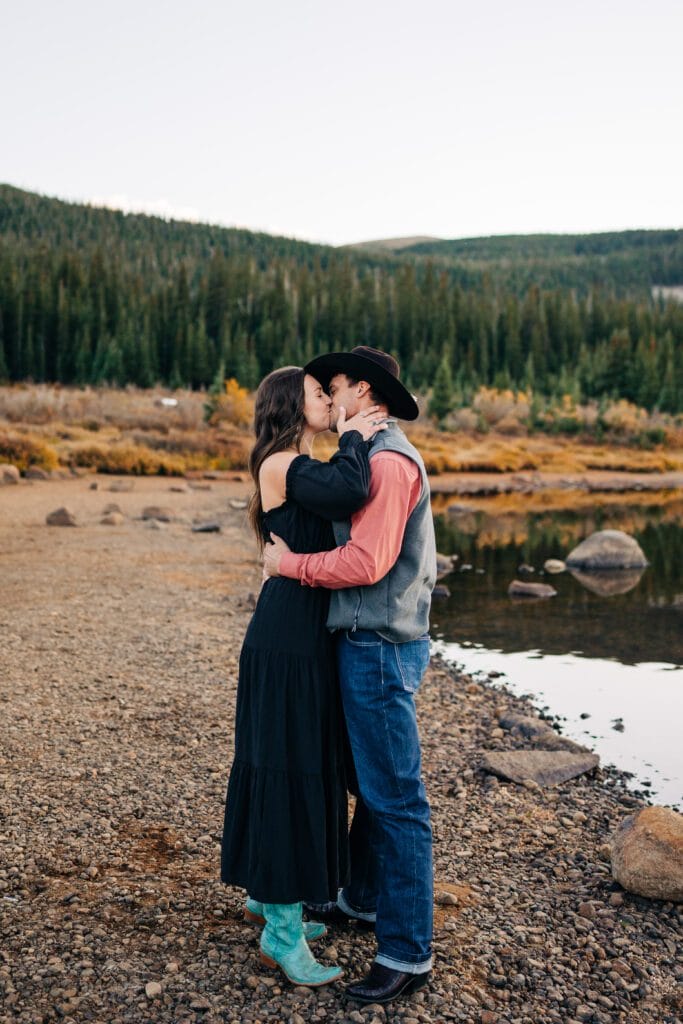A couple kisses while standing on a rocky lakeshore, surrounded by trees and distant hills. The woman wears a black dress and turquoise boots, while the man wears a cowboy hat, jeans, and a vest.
