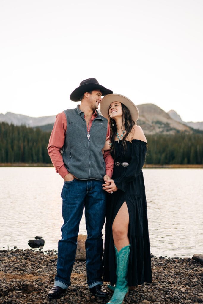 A man and woman in western attire stand by a lake, smiling at each other. The man wears a cowboy hat, vest, and jeans; the woman wears a hat, off-shoulder black dress, and turquoise boots. Mountains and trees are in the background.