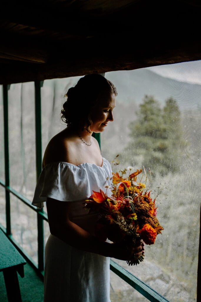 A woman in an off-shoulder white dress stands by a window, holding a bouquet of autumn flowers, looking down thoughtfully. Trees and mountains are visible outside through the large window.