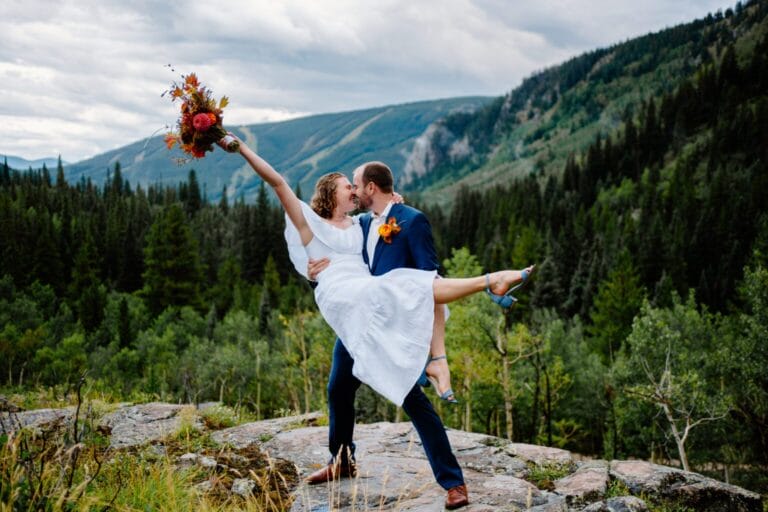 A groom in a blue suit lifts a bride in a white dress as she holds a bouquet aloft. They smile joyfully on a rocky ledge with green forest and mountains in the background under a cloudy sky.