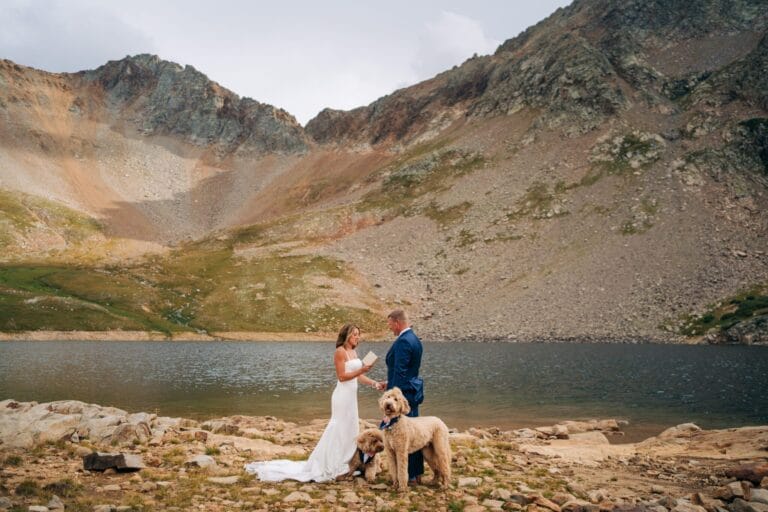 A bride and groom stand beside a mountain lake during their wedding ceremony, holding hands with two dogs on leashes in front of them, surrounded by rocky terrain and tall peaks.