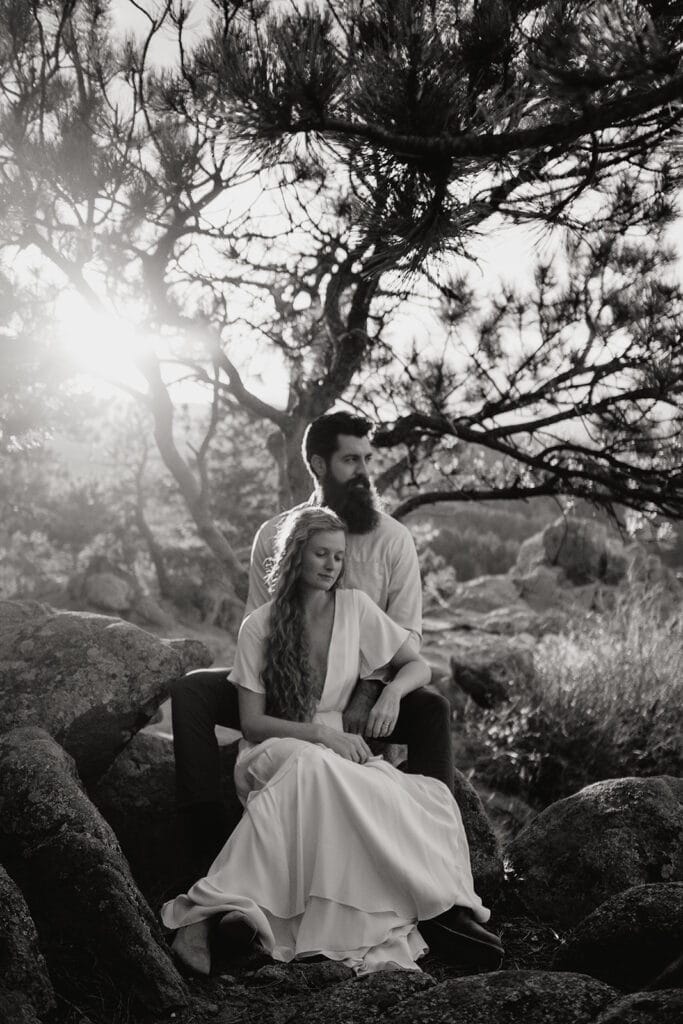 A couple sits together on rocks under a tree, bathed in sunlight. The woman in a flowing dress sits in front, leaning back against the man, who has a beard. Both appear calm and serene in the natural setting. Black and white photo.