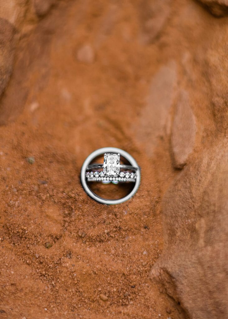 A pair of silver rings, one with a rectangular diamond and the other with small round stones, arranged together on reddish-brown, sandy ground.