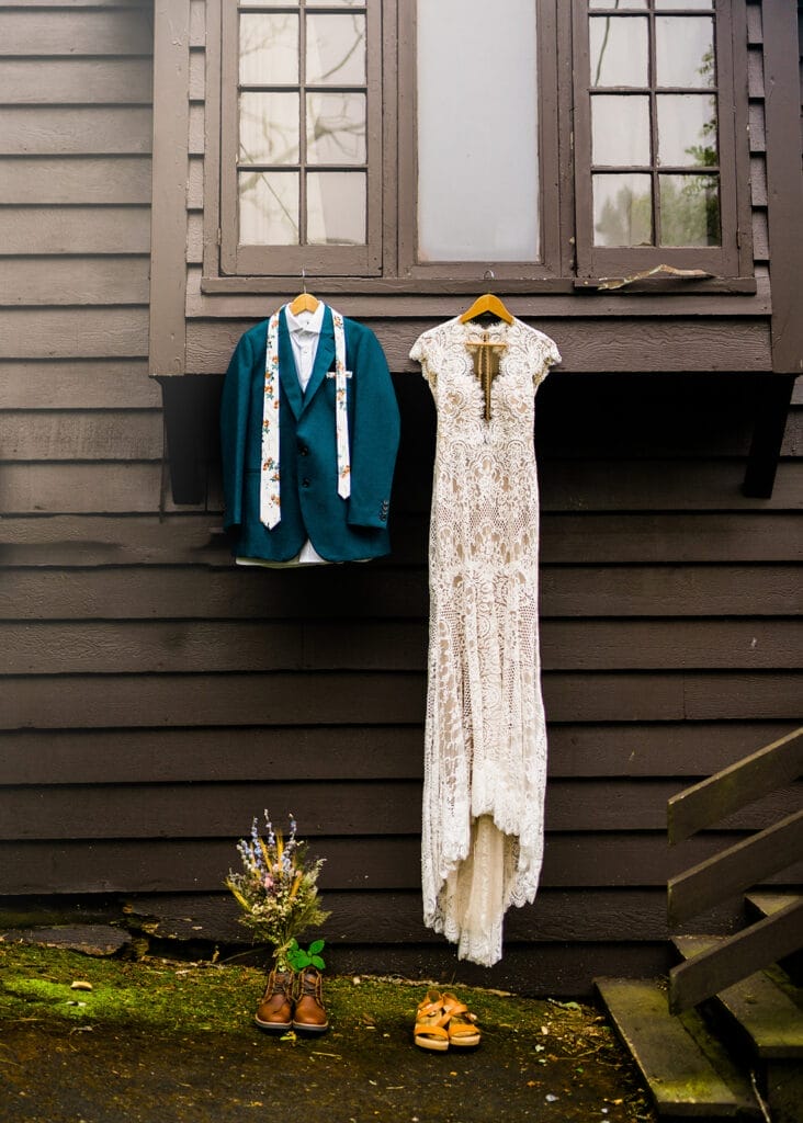 A teal jacket with a floral tie and a white lace wedding dress hang on a brown wooden wall beneath windows. Below them are a bouquet, brown shoes, and tan sandals on the ground.
