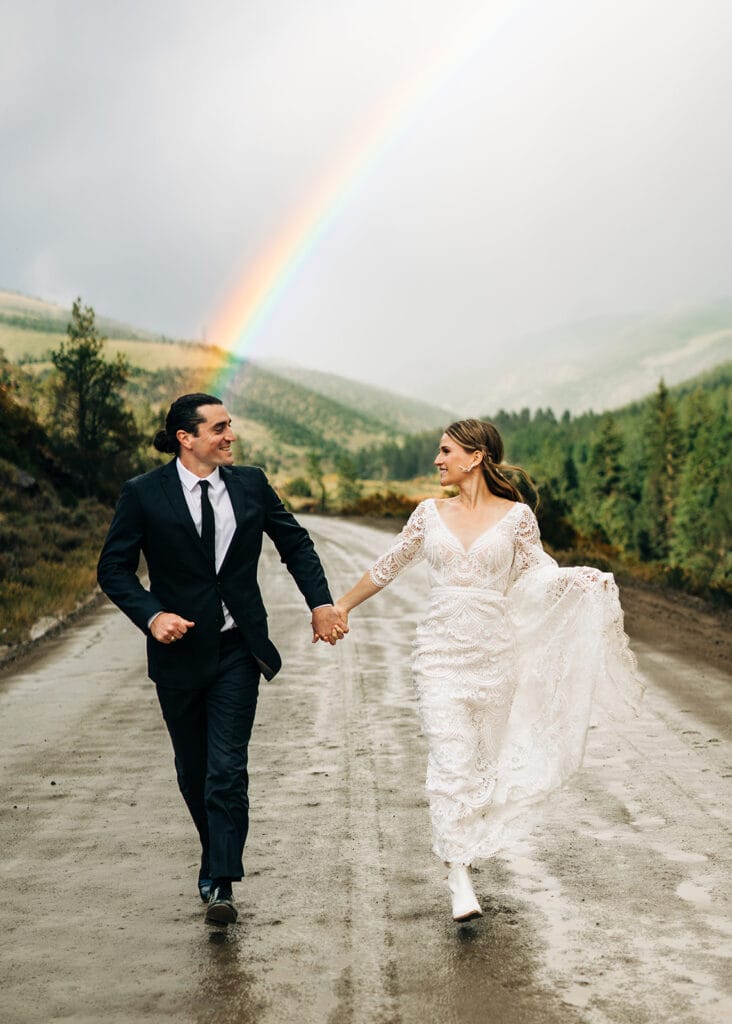 A bride and groom hold hands and walk joyfully down a wet road surrounded by greenery, with a vivid rainbow arching over the hills in the background.