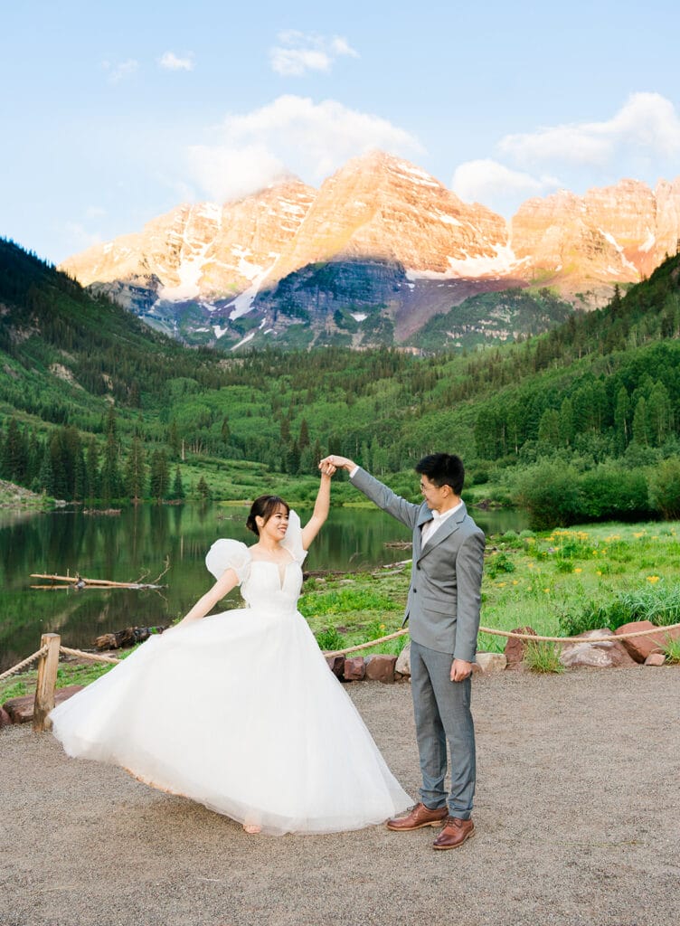 A bride in a white dress twirls joyfully with a groom in a gray suit, set against lush green mountains and a lake, with snow-capped peaks glowing in the background.